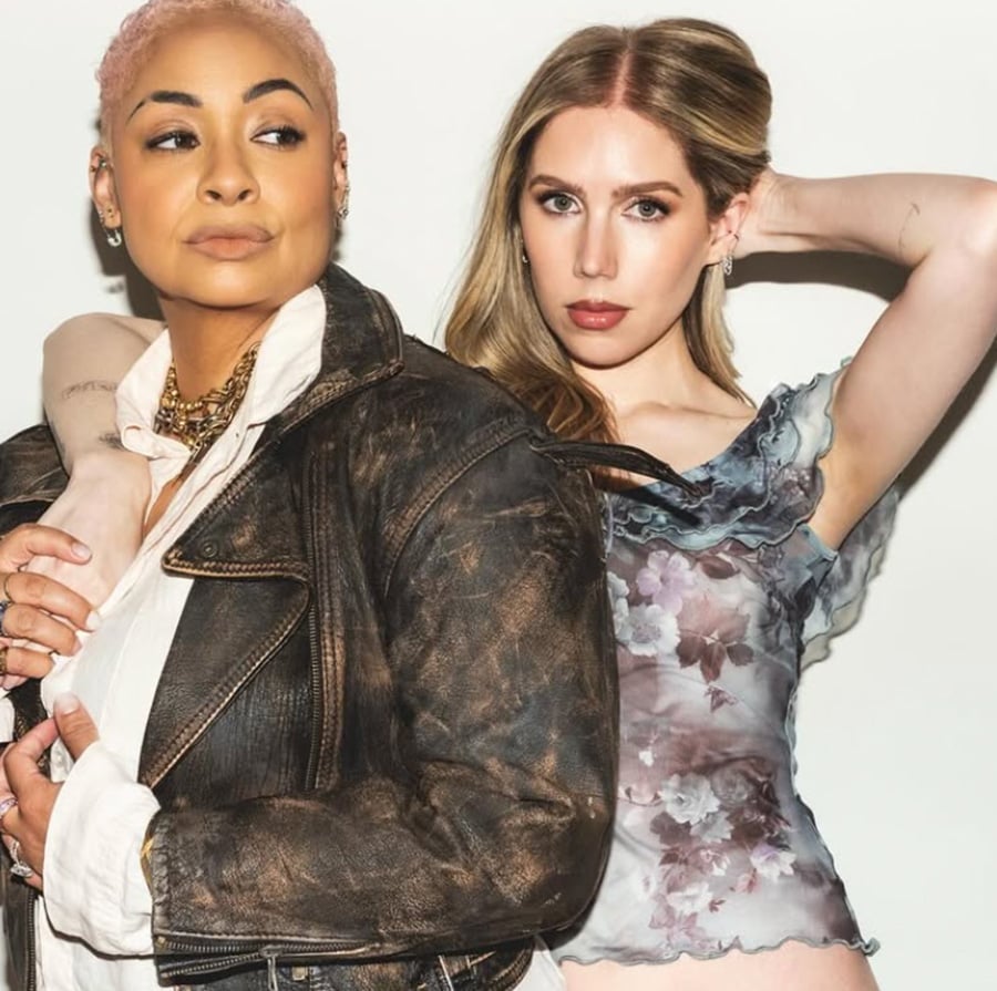 Two women pose confidently against a white background. One wears a black leather jacket over a white shirt with layered necklaces, while the other has her arm around her, wearing a sheer floral top and holding her hair back.