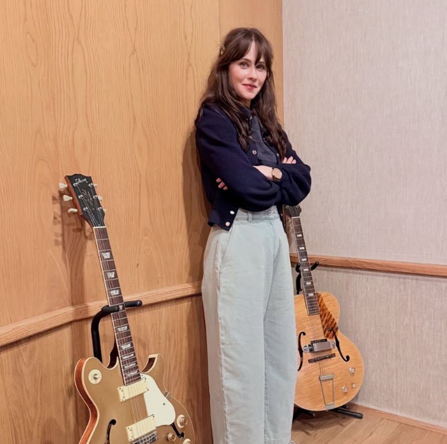 A woman with long brown hair stands with arms crossed, smiling between two electric guitars on stands, against a wall with wood paneling and beige wallpaper.
