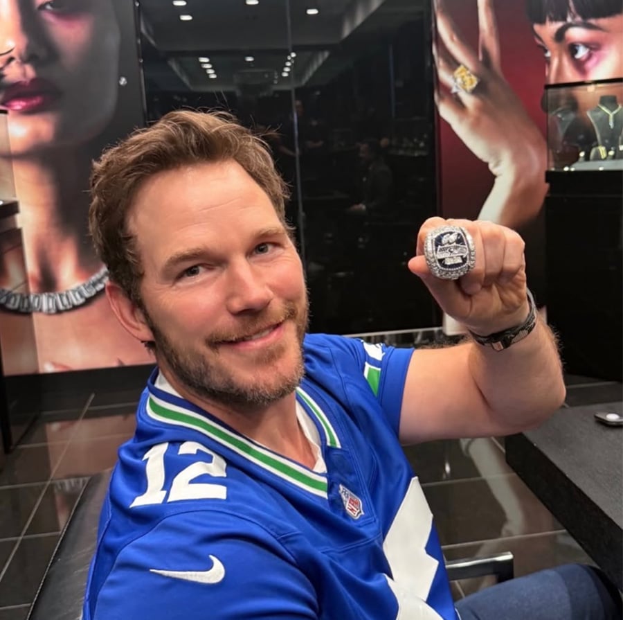 A smiling man wearing a blue sports jersey with the number 12 holds up a large ring in a jewelry store. The background features wall art of models wearing jewelry.
