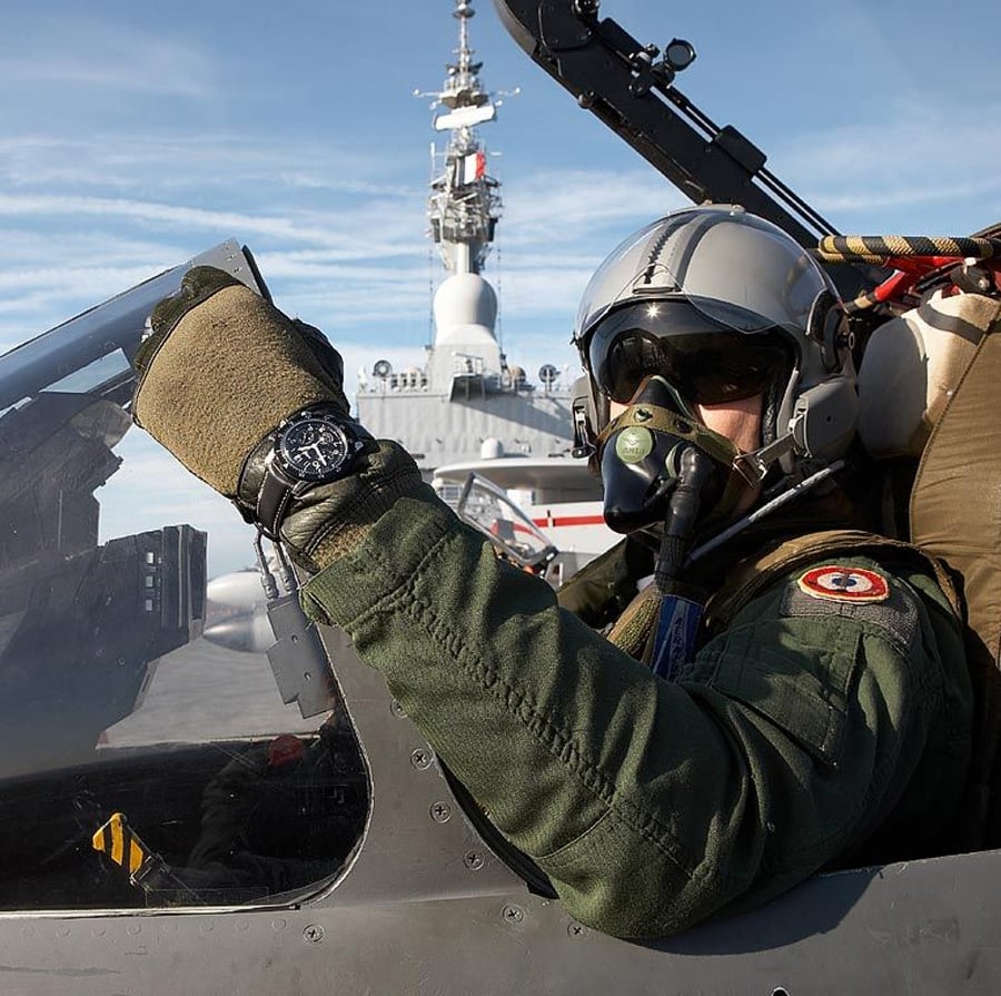 A fighter pilot wearing a helmet, oxygen mask, and green flight suit sits in a jet cockpit, giving a thumbs-up. A naval ship is visible in the background under a blue sky.
