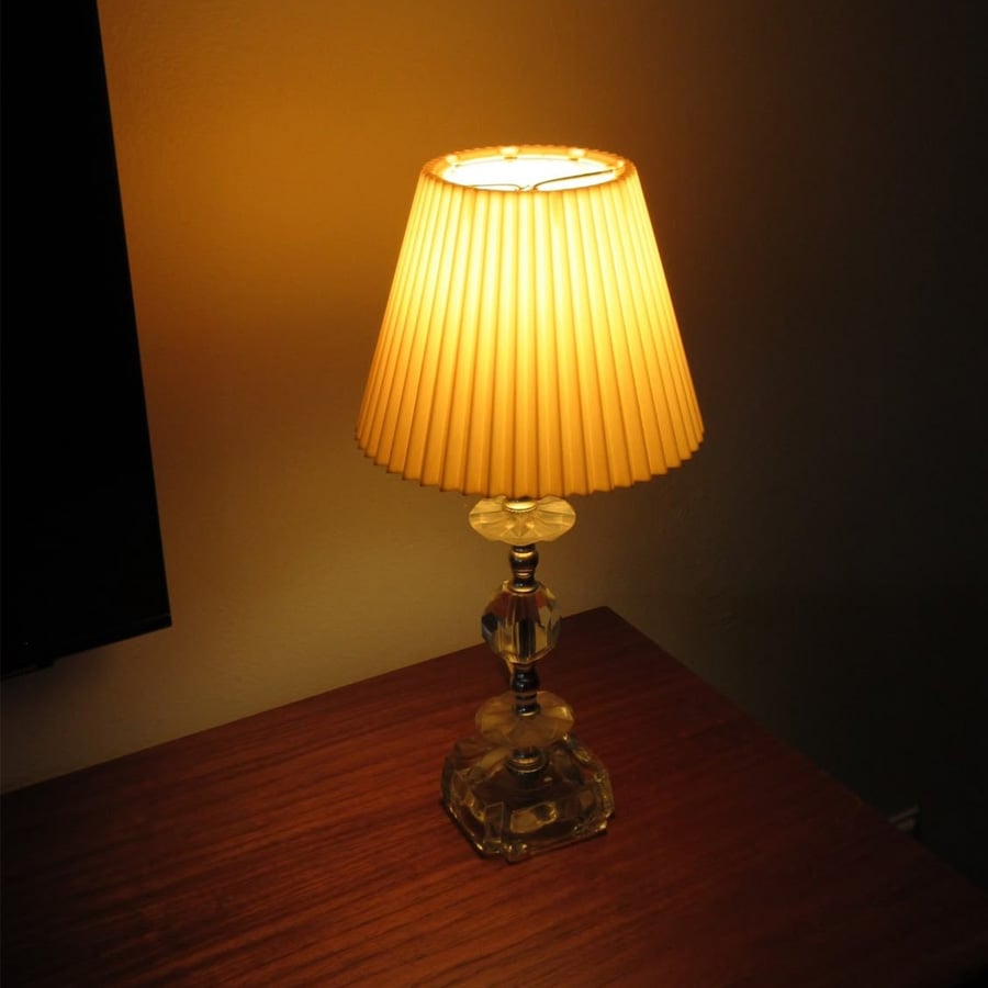 A small table lamp with a pleated beige lampshade casts a warm yellow glow on a wooden surface in a dimly lit room.