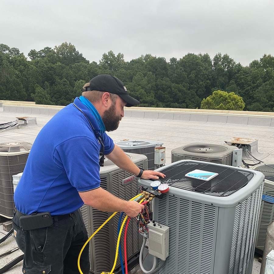 A technician in a blue shirt and cap works on rooftop HVAC units, using gauges and colored hoses to check the equipment, with trees visible in the background under a cloudy sky.