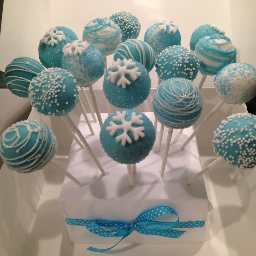 Blue and white cake pops decorated with sprinkles, snowflakes, and swirled icing are arranged upright on white sticks in a white stand with a blue ribbon bow.