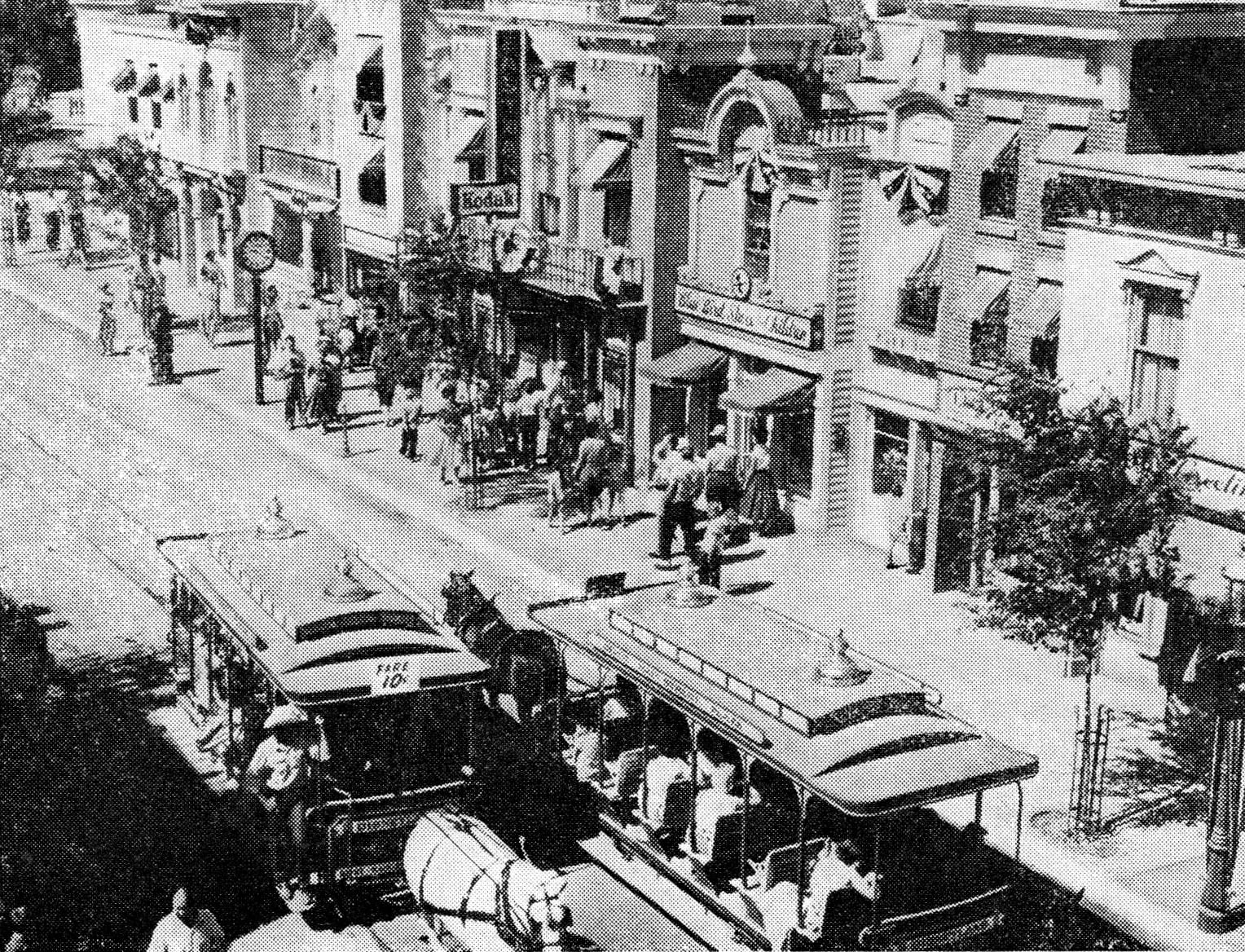 A black-and-white photo shows a lively street scene with horse-drawn trolley cars, people walking on the sidewalk, and storefronts including a Kodak shop and a theater.