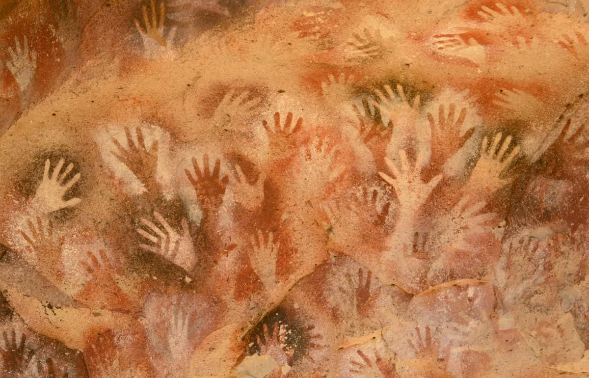 Cave wall covered with numerous handprints, mostly in white and reddish-brown hues; the hand shapes overlap and vary in size, creating a densely patterned ancient artwork.