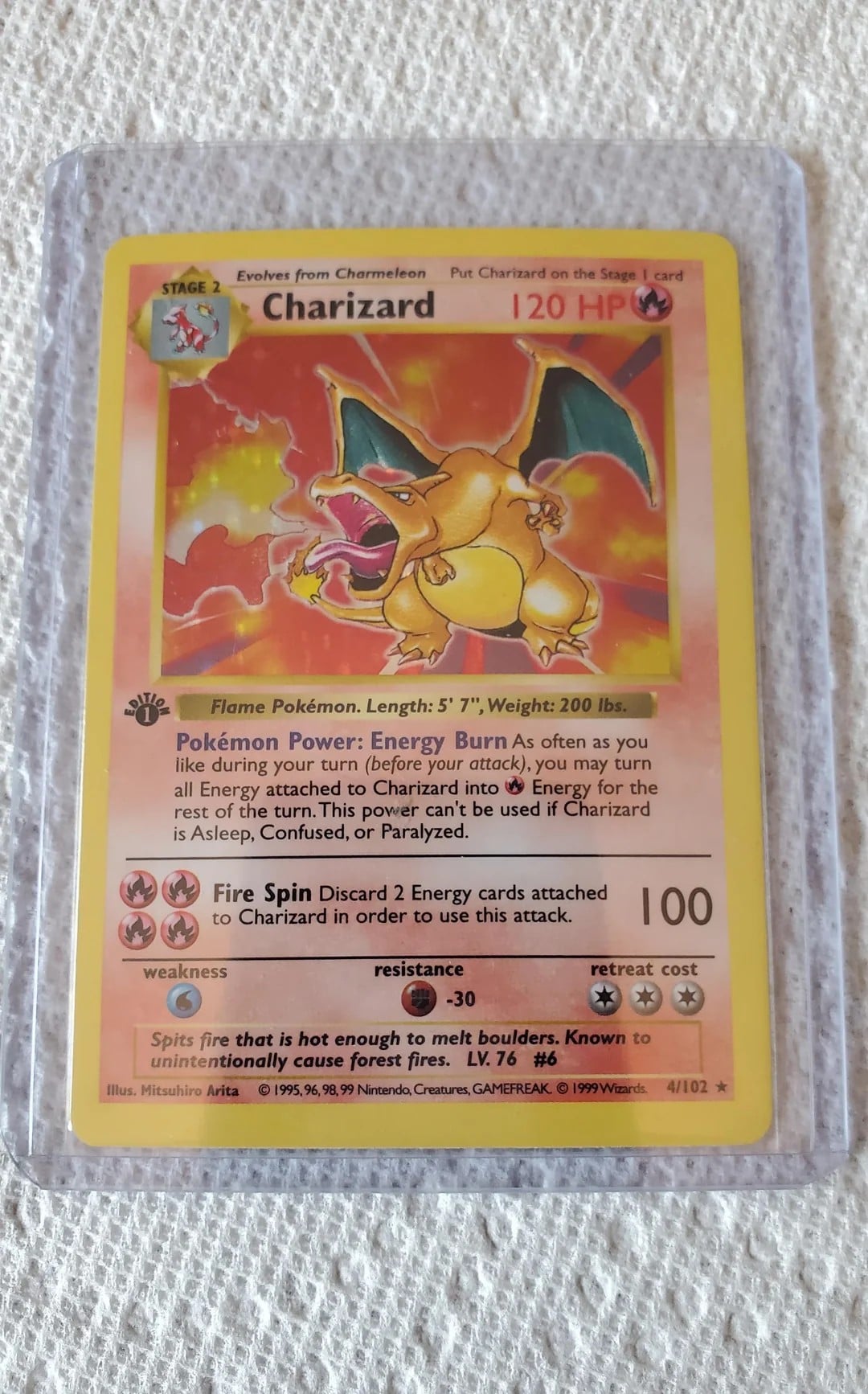 A Charizard Pokémon trading card in a plastic sleeve is placed on a textured white surface. The card features an illustration of Charizard breathing fire, with stats and abilities visible.