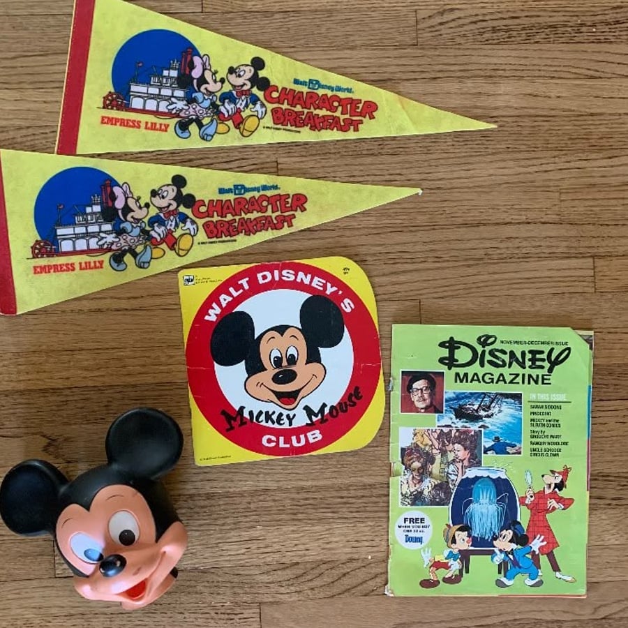 A collection of vintage Disney memorabilia, including two yellow Character Breakfast pennants, a Mickey Mouse Club sign, a Disney Magazine, and a Mickey Mouse face toy, all arranged on a wooden surface.