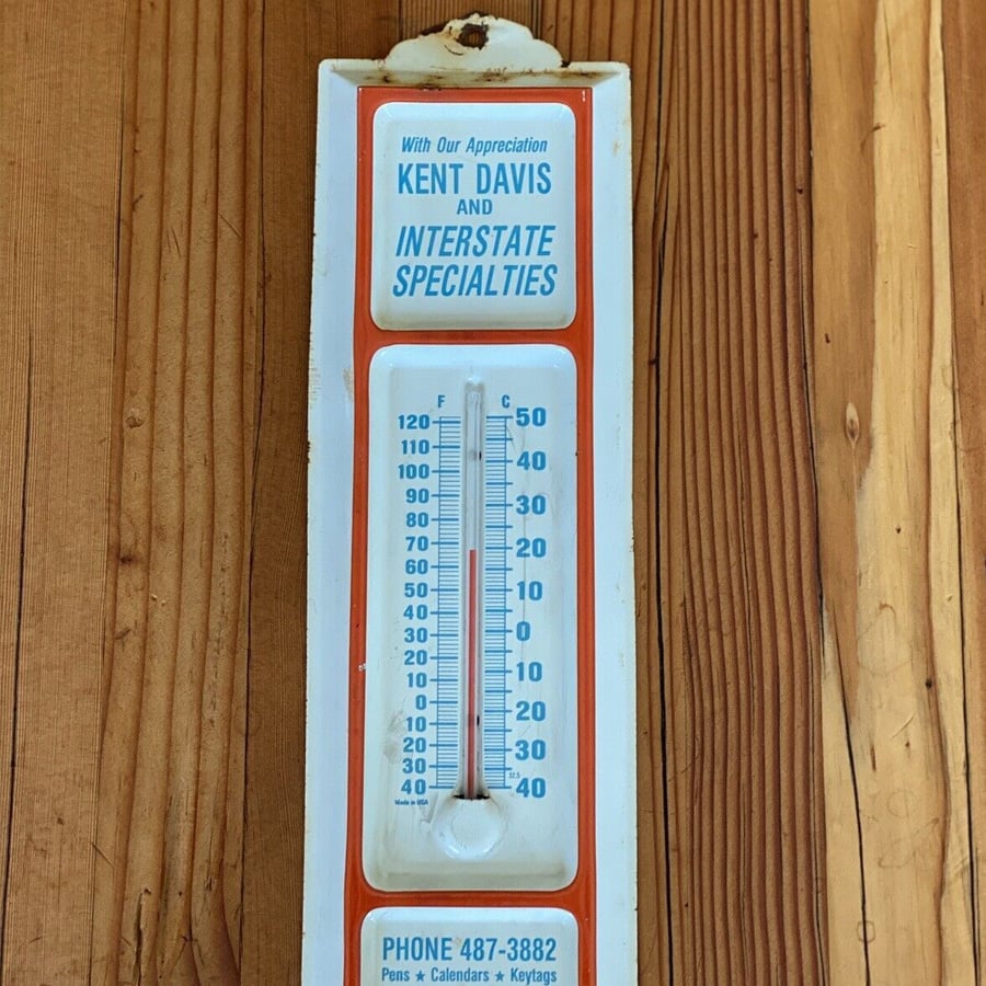 A vintage wall thermometer with blue text reading "Kent Davis and Interstate Specialties" and a phone number, mounted on wood paneling, showing a temperature of about 74°F.