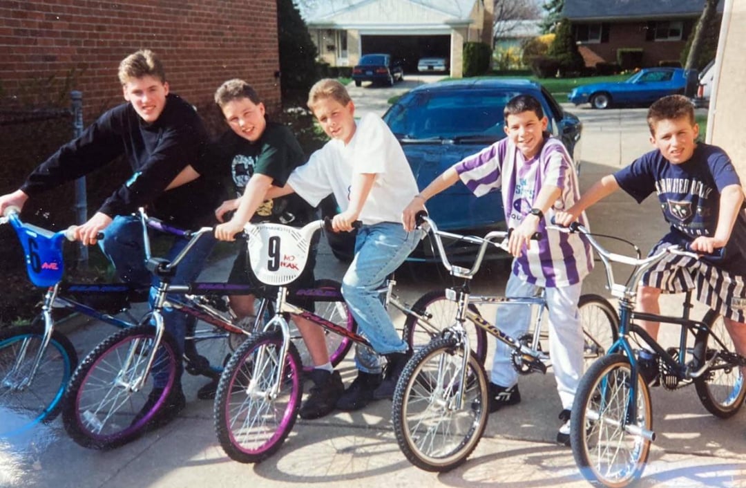 Five boys pose together outside on bicycles in a driveway, smiling at the camera. Houses and parked cars are visible in the background on a sunny day.