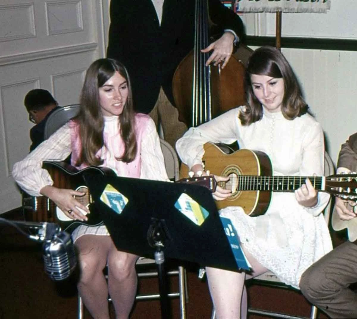 Two young women sit side by side playing acoustic guitars, reading sheet music on a stand. Both wear light-colored dresses and are performing indoors, with a microphone visible in the foreground.
