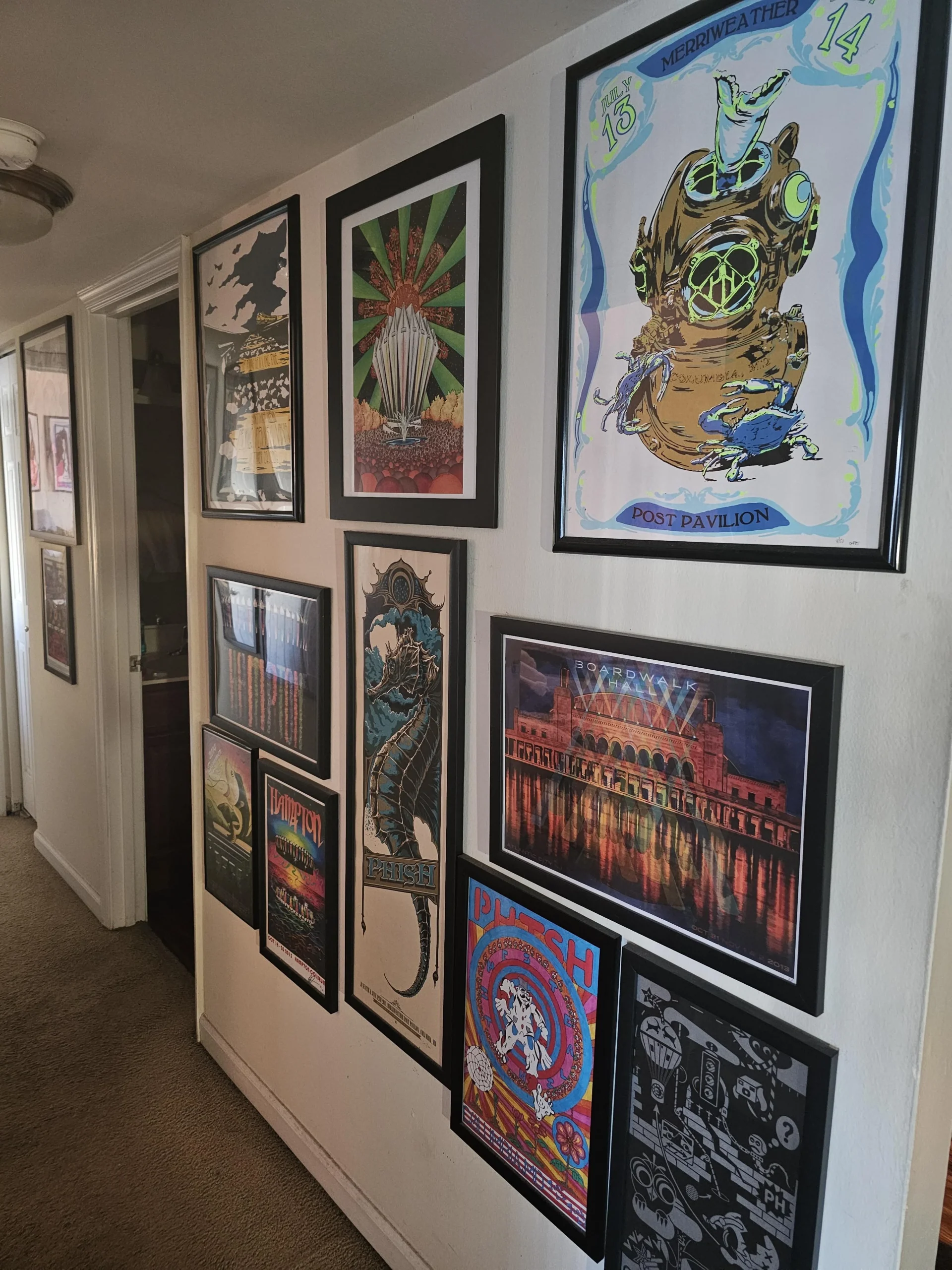 A hallway wall displaying a colorful gallery of framed concert posters, featuring various artistic designs including sea creatures, circus-themed art, and a diving suit illustration. The floor is carpeted and doors line the hallway.