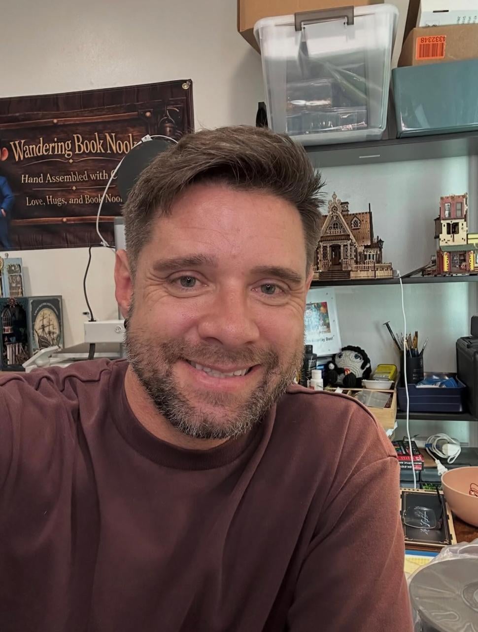 A man with short brown hair and a beard smiles at the camera in a room filled with books, shelves, and intricate miniature buildings behind him.