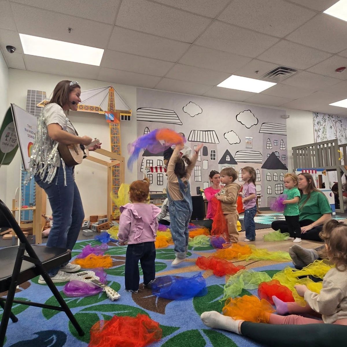 A woman plays guitar and sings to a group of young children in a colorful playroom. The children wave bright scarves and dance, while adults sit nearby, watching and engaging. The room has playful wall art with houses and clouds.