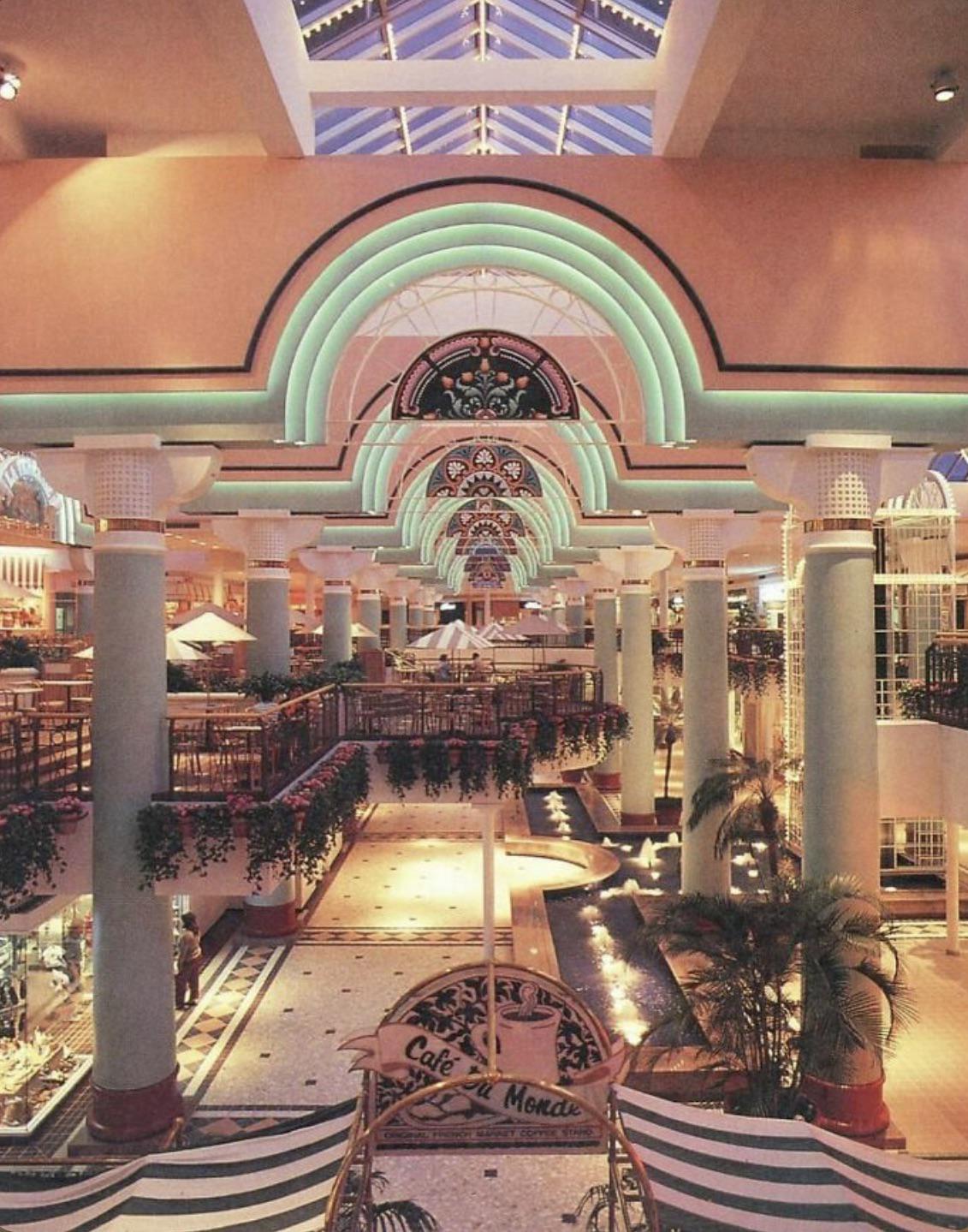 A spacious, colorful shopping mall interior with pastel arches, tall columns, skylights, hanging plants, and decorative lighting. Café tables and greenery are visible, creating an inviting, retro atmosphere.