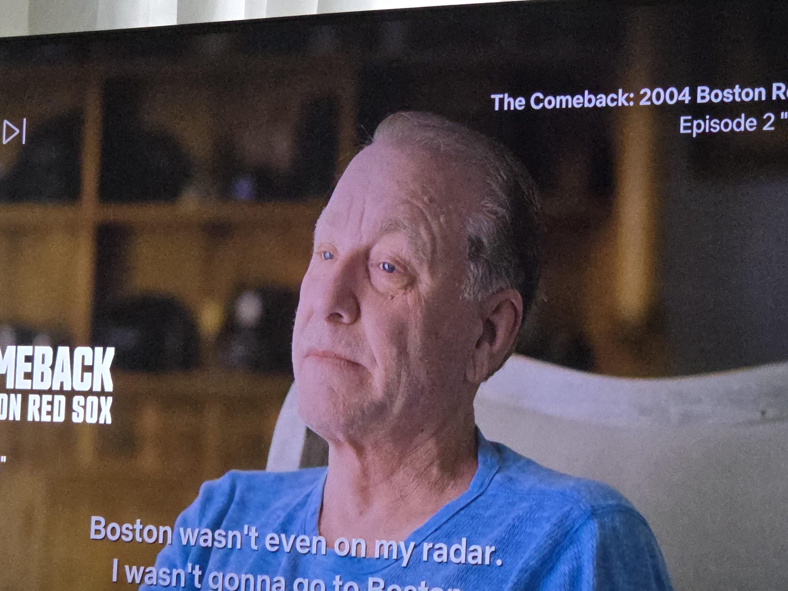 A man with short, light hair wearing a blue shirt sits indoors, looking serious. White text on the screen references "The Comeback: 2004 Boston Red Sox" and partial subtitles appear at the bottom.