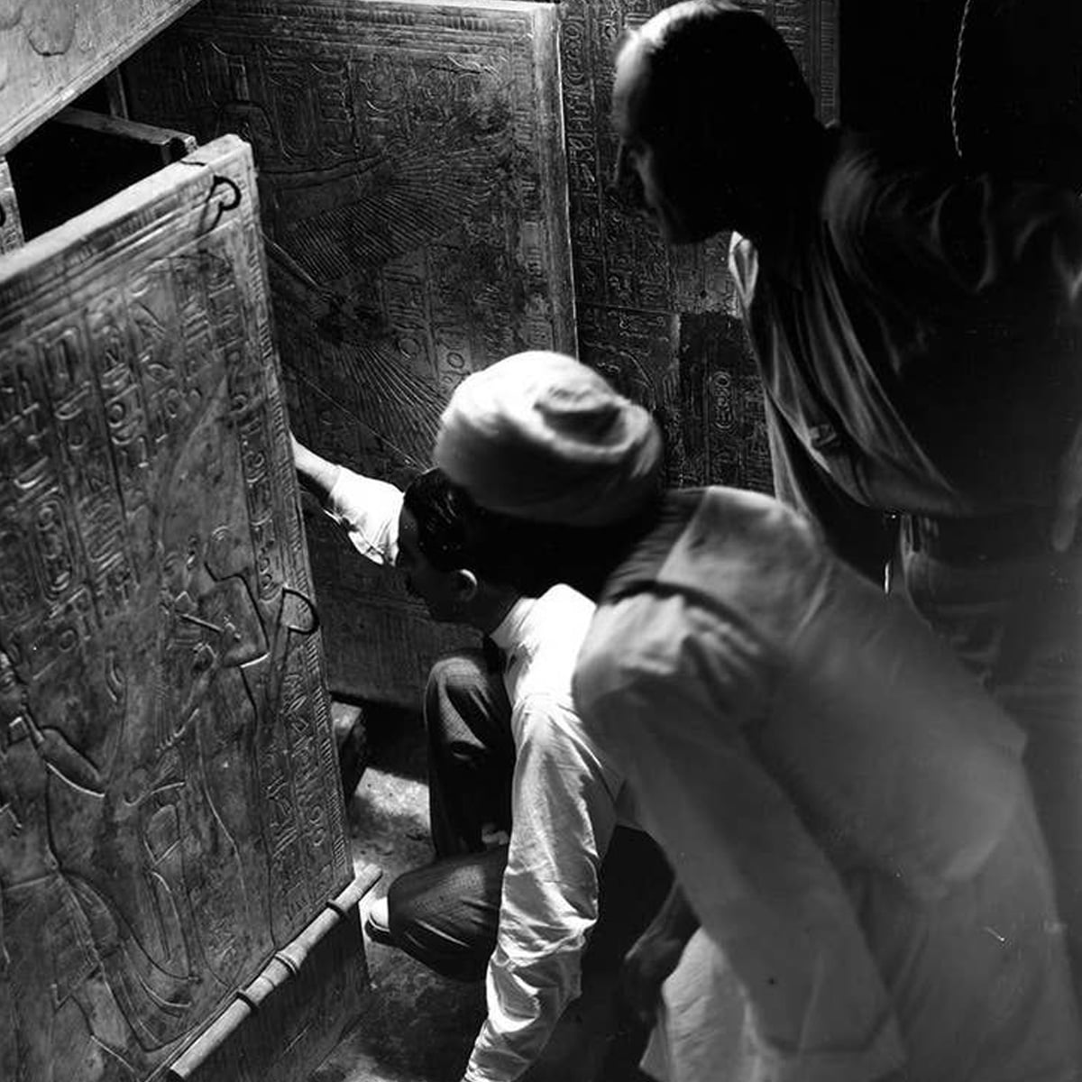 Three men closely examine ancient Egyptian hieroglyphs carved on large stone panels in a dimly lit room, with one man kneeling and pointing at the carvings.