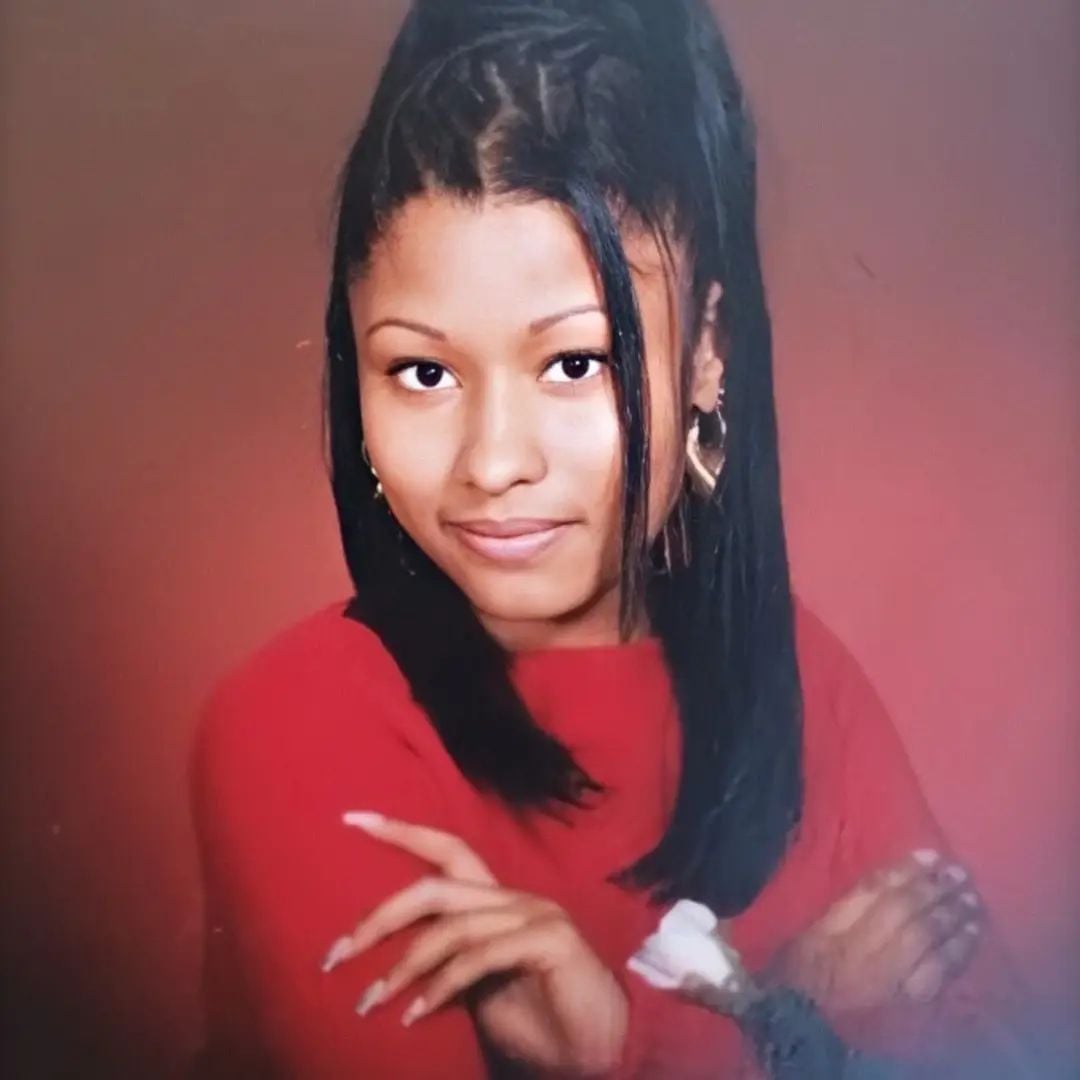 A young woman with long, dark hair styled in a half-up ponytail poses against a reddish-brown background. She is wearing a red top, gold hoop earrings, and has her arms crossed with a slight smile.