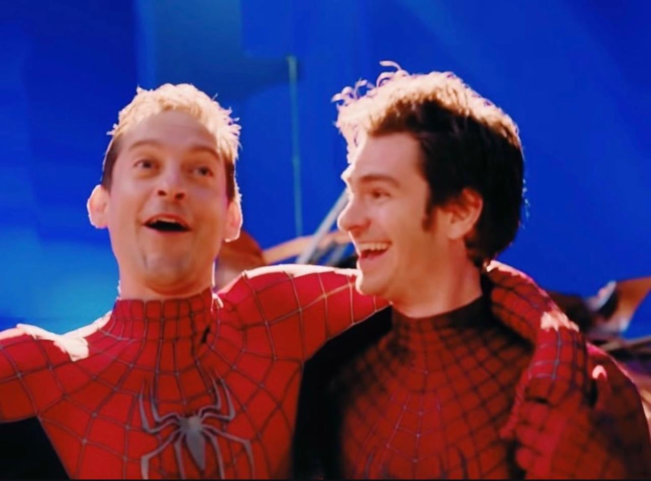 Two people dressed in Spider-Man costumes stand side by side with their arms around each other, smiling and laughing against a blue background.