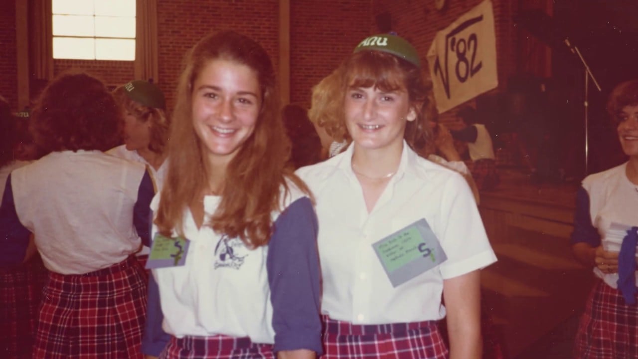 Two teenage girls wearing plaid skirts and white shirts stand smiling indoors at what appears to be a school event. Other students in similar outfits and a sign reading “‘82” are visible in the background.