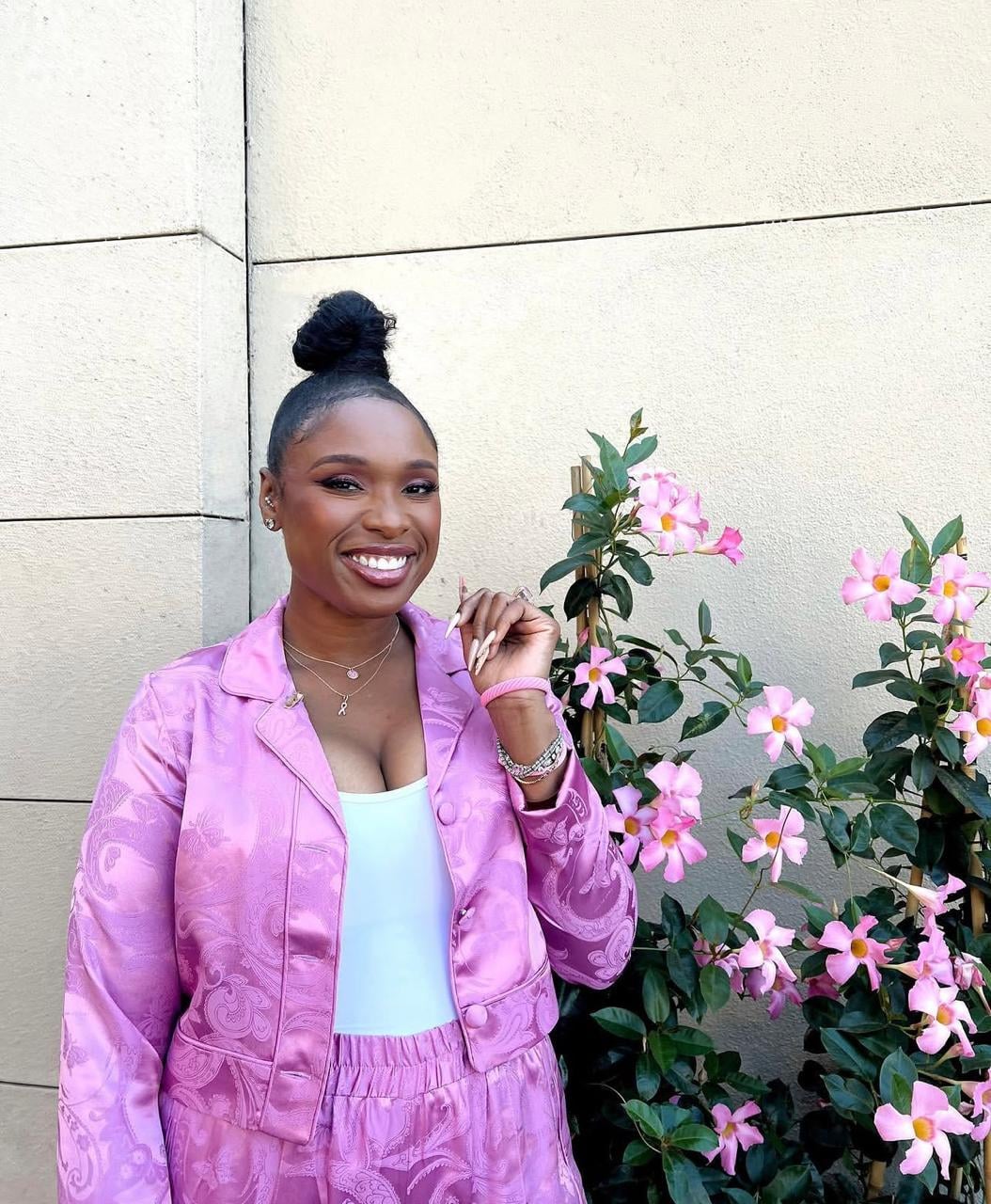 A woman in a pink patterned jacket and pants stands smiling beside a flowering bush with pink blossoms, against a light-colored wall. She wears her hair in a top bun and has a white top underneath her jacket.