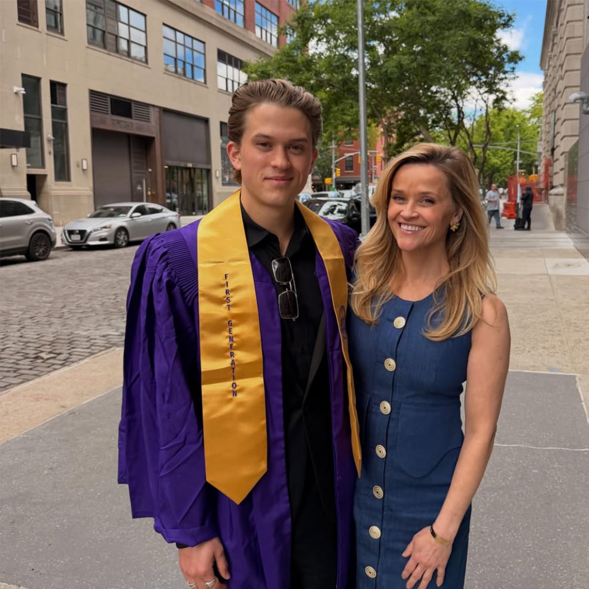 A young man in a purple graduation gown with a gold stole stands on a city sidewalk next to a smiling woman in a sleeveless blue dress. Trees and buildings line the street in the background.