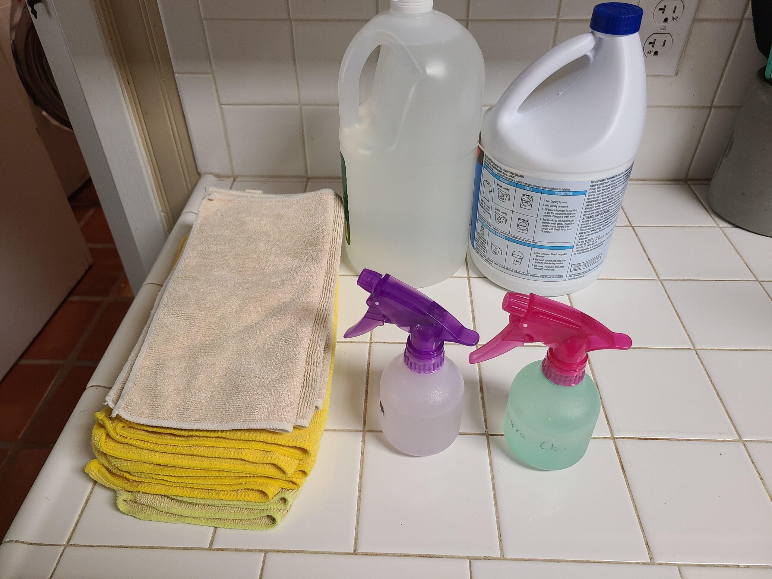 Cleaning supplies on a white tiled counter: a stack of yellow cloths, a clear gallon jug, a white bleach bottle, and two small spray bottles, one purple and one pink, filled with liquid.