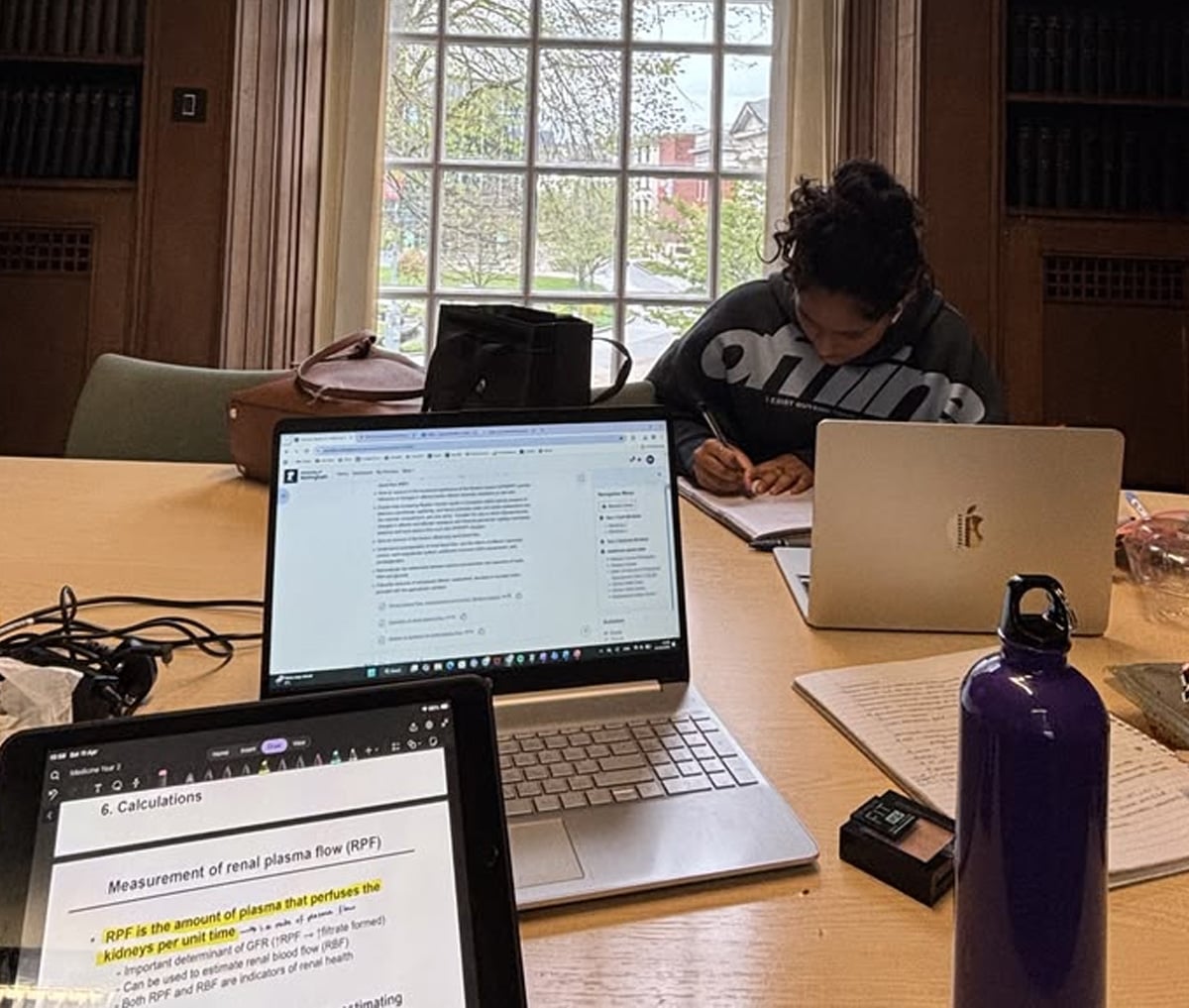 A study room with three laptops on a wooden table, notebooks, a water bottle, and a person writing. Sunlight streams through a large window in the background, showing trees and a building outside.