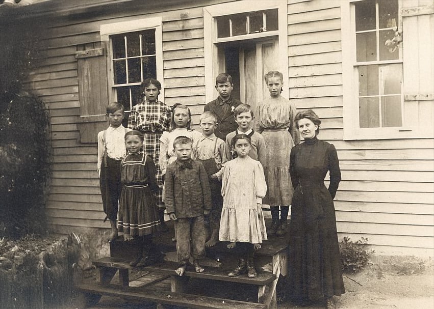 A vintage photo shows a group of children and a woman standing on wooden steps outside a wooden building with windows. The children wear old-fashioned clothes, and the woman stands to the right, likely a teacher.