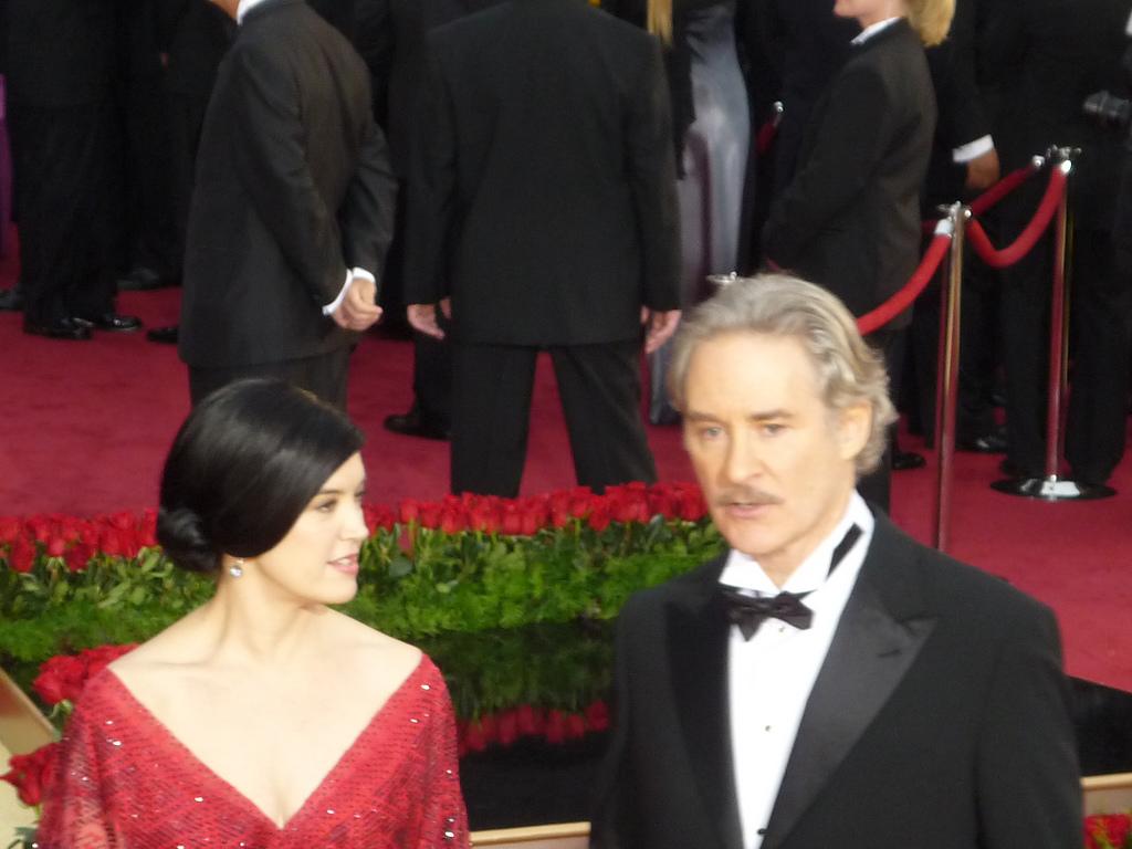 A woman in a sparkling red dress and a man in a black tuxedo stand on a red carpet, surrounded by people in formal attire. Red ropes and flower arrangements are visible in the background.