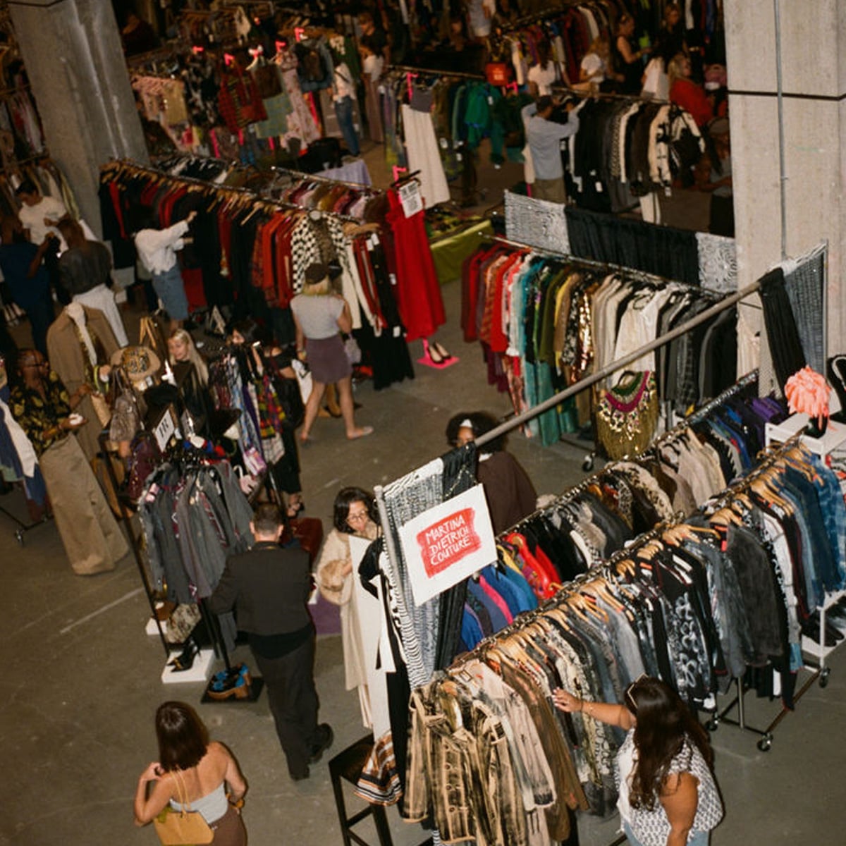 A busy indoor vintage clothing market with racks of colorful clothes, shoppers browsing, and signs marking different sections. Some people are inspecting garments while others walk between the aisles.
