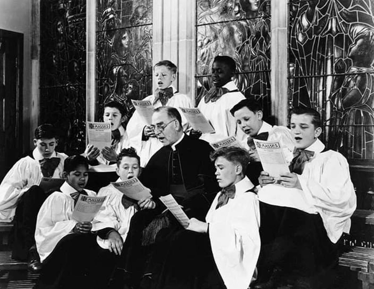 A group of choirboys and a clergyman sit together, dressed in choir robes, holding music booklets and singing in front of ornate stained-glass windows.