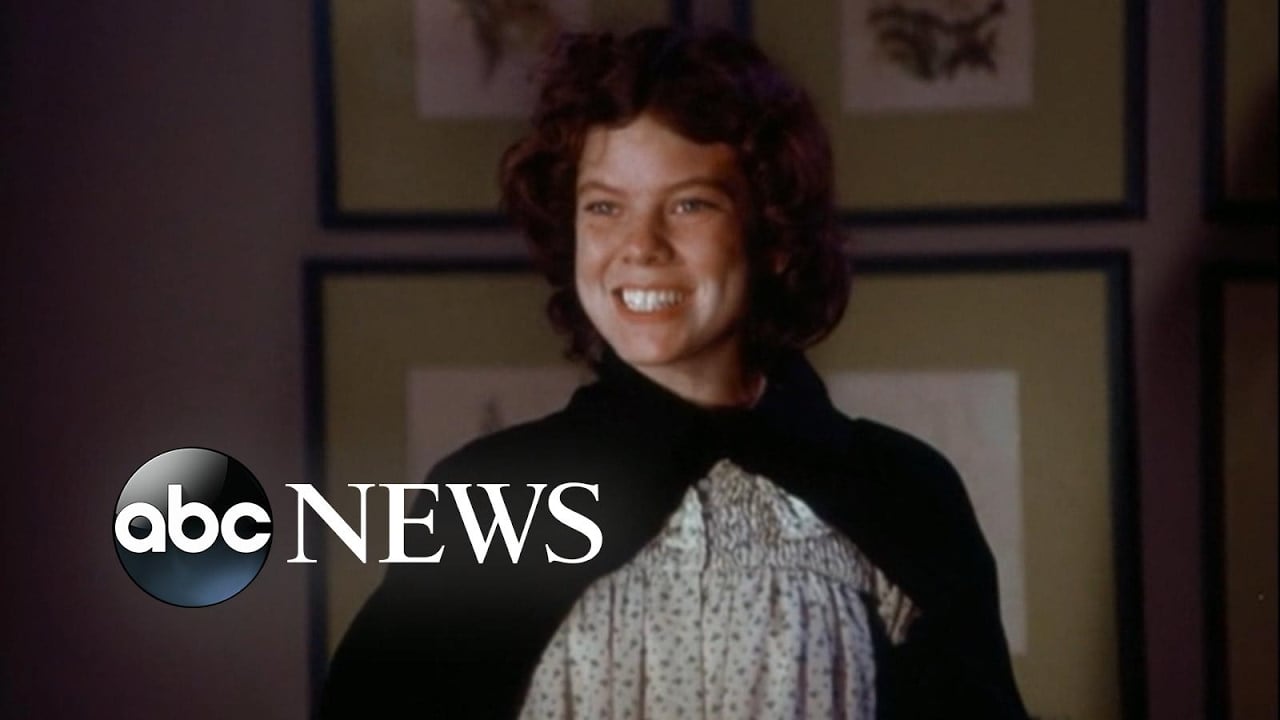A young person with curly hair smiles widely while wearing a cloak over a light-colored dress. The ABC News logo is overlaid in the lower left corner of the image. Framed pictures hang on the wall in the background.