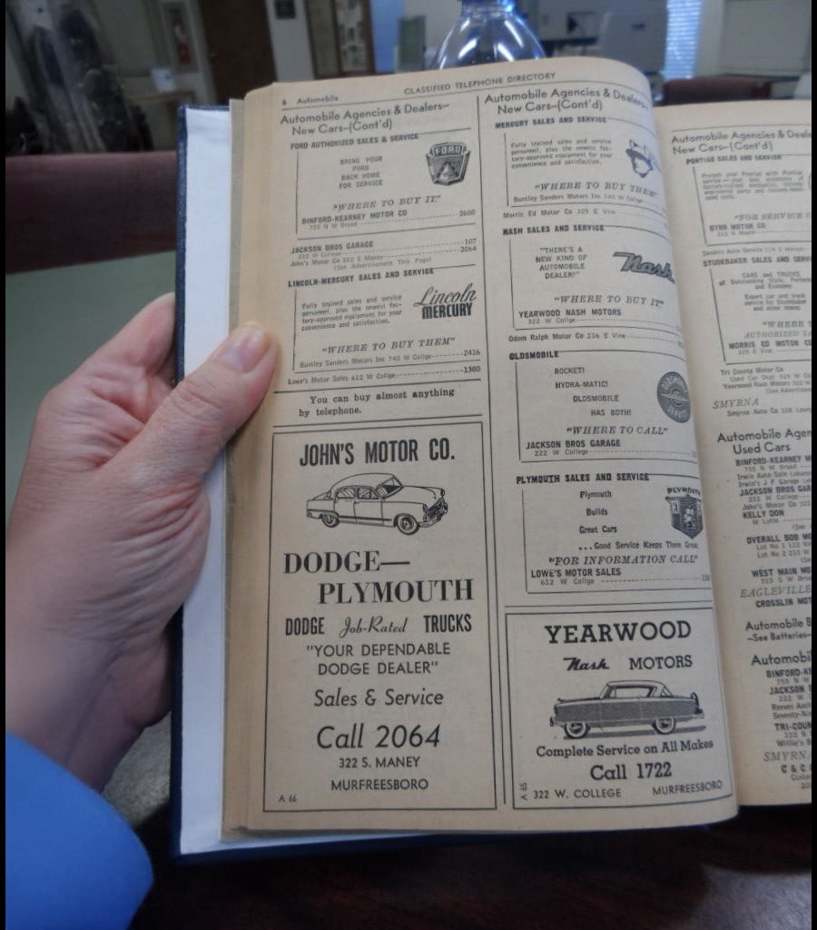 A hand holds open a vintage yellow pages directory showing ads for car dealerships, including Dodge, Plymouth, Mercury, and StudeBaker, with phone numbers and addresses in Murfreesboro, Tennessee.