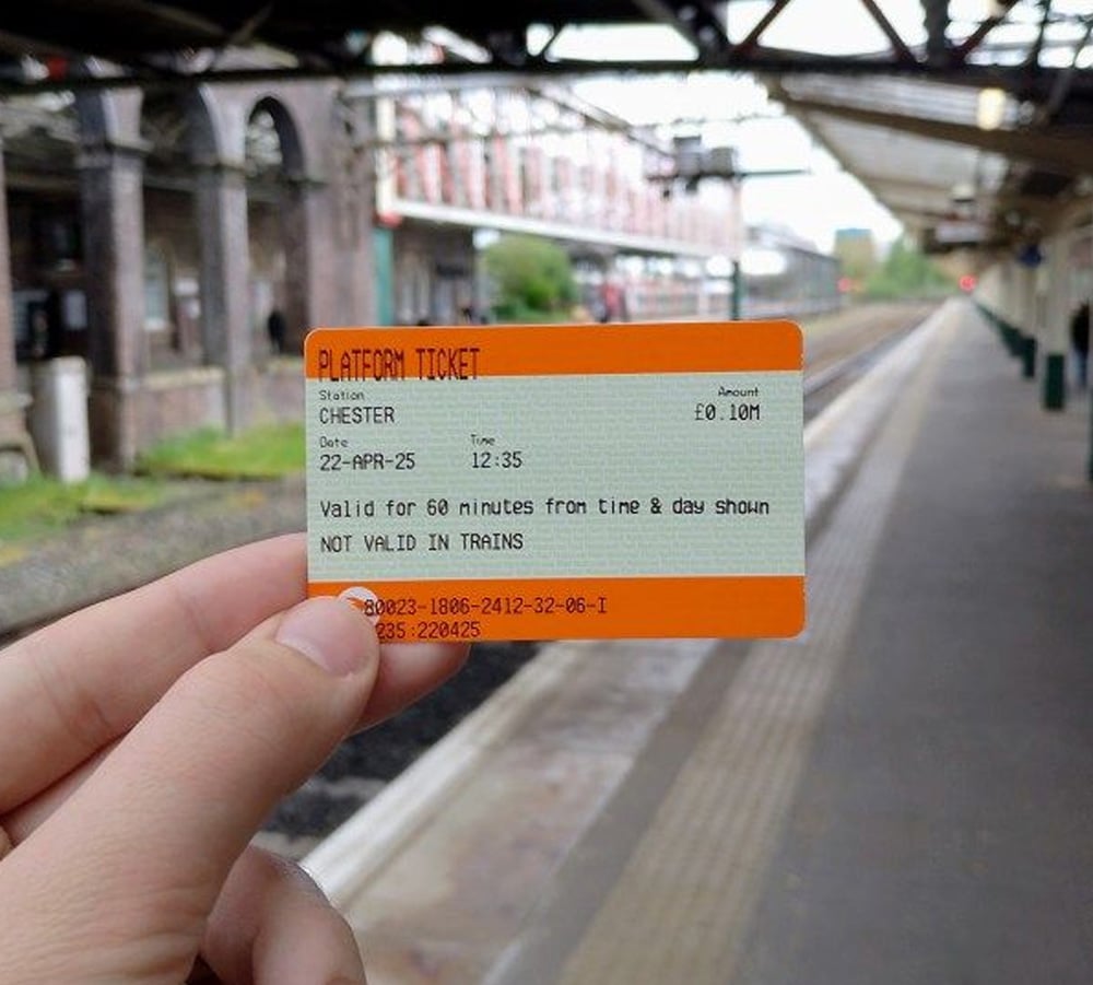 A hand holds a platform ticket at a train station. The ticket is orange and white, showing details such as "Chester," the date, time, and validity, with empty tracks and a station platform in the background.