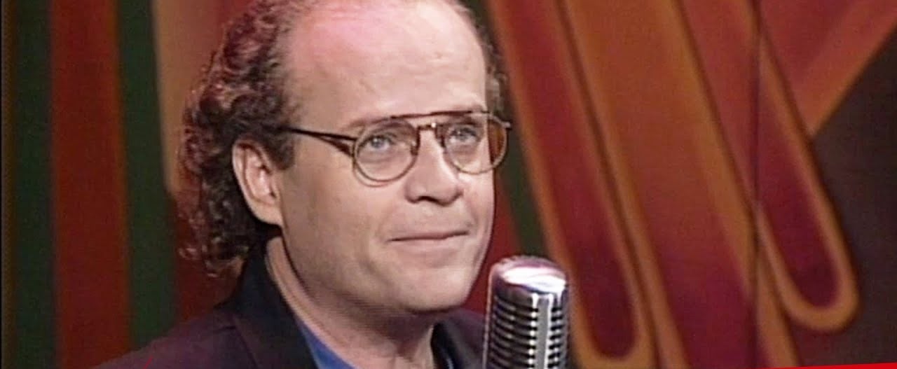 A man with glasses, receding curly hair, and a serious expression stands in front of a retro microphone and colorful abstract background.