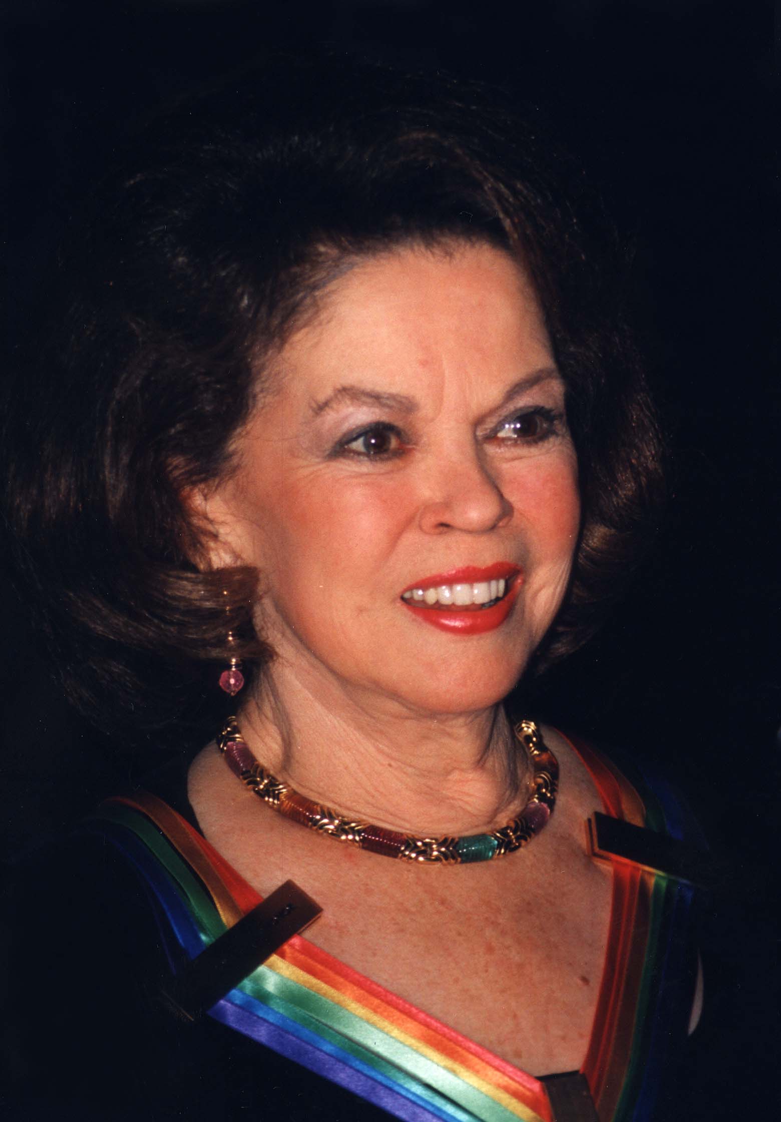 A smiling woman with short brown hair wears a dark dress with a rainbow-colored collar, gold necklace, and earrings, against a dark background.