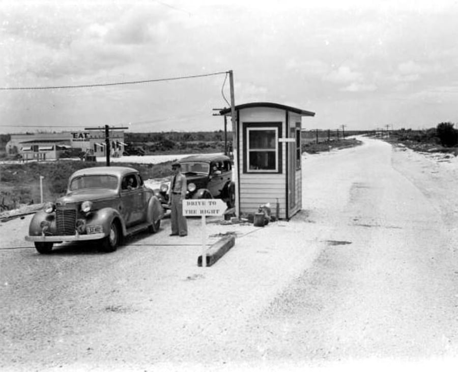 Black-and-white photo of vintage cars stopped at a small roadside booth. A person stands by the booth, and a sign reads “Drive to the right.” A diner sign reading “EAT” is visible in the background.