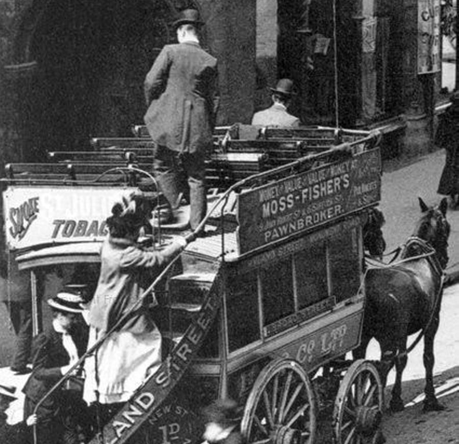 A black-and-white photo of people boarding a double-decker horse-drawn carriage on a city street. The carriage, advertising a pawnbroker, is pulled by two horses, and several passengers wear hats and coats.