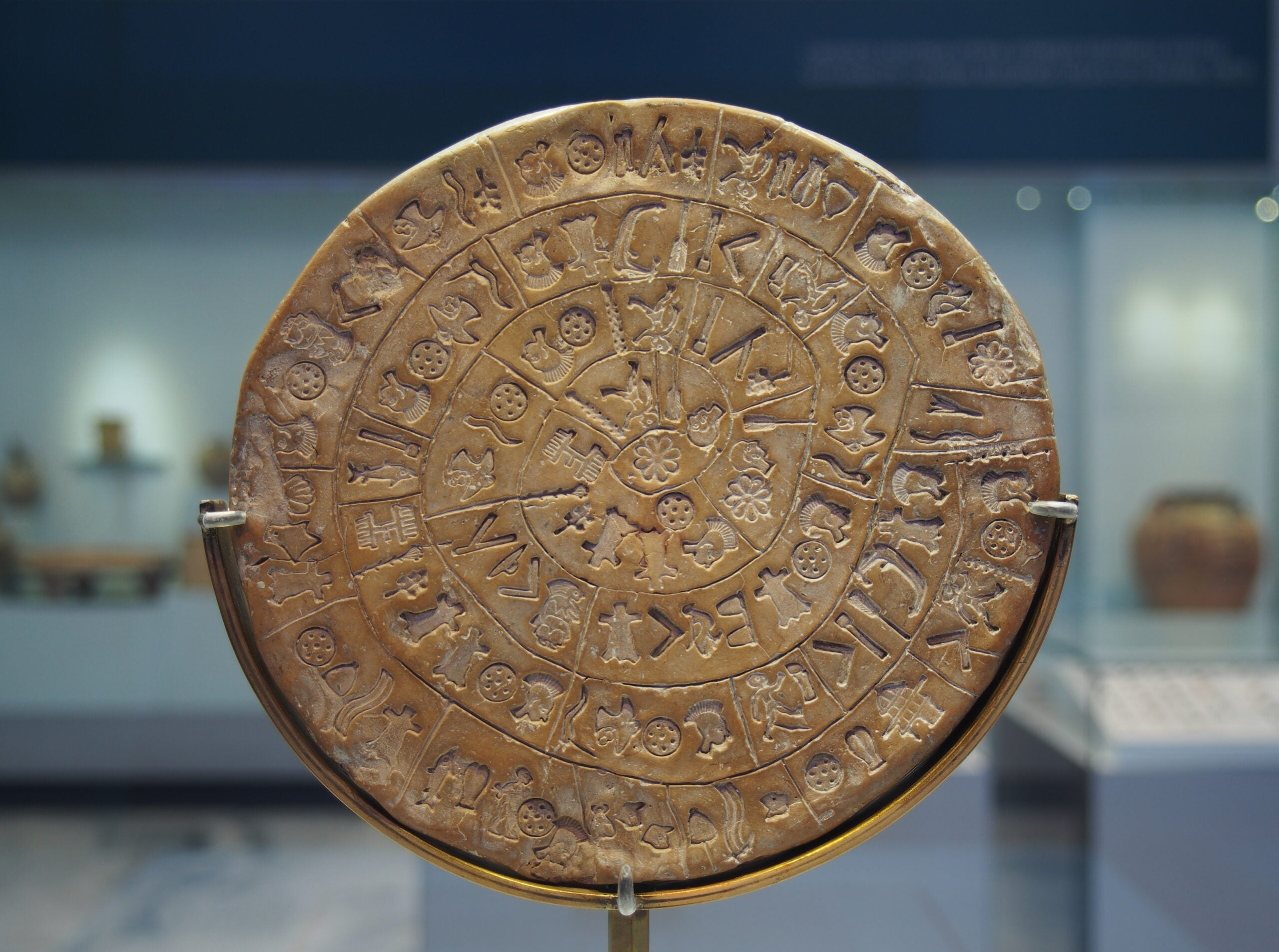 A close-up of the Phaistos Disc, a round clay artifact inscribed with spiral rows of mysterious symbols and pictograms, displayed upright in a museum exhibit.