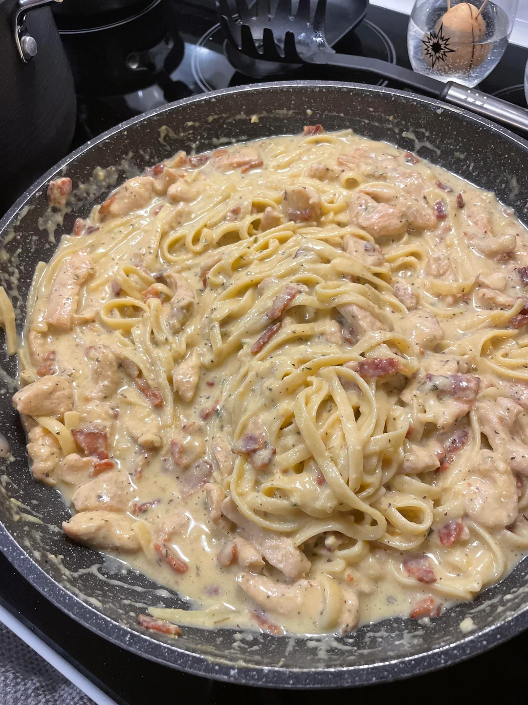 A skillet filled with creamy pasta, likely fettuccine, mixed with pieces of chicken and bacon in a rich sauce, sitting on a stovetop with kitchen utensils and a pot visible in the background.