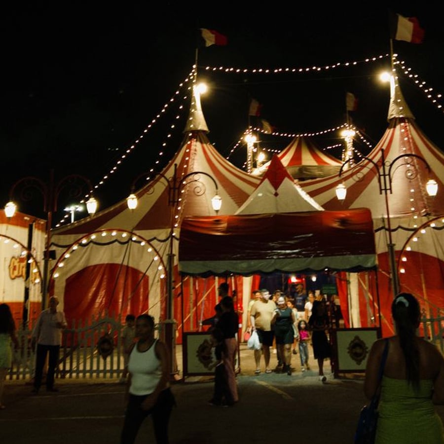 A brightly lit circus tent with red and white stripes is seen at night. People are entering and exiting through the entrance, and strings of lights and flags decorate the top of the tent.