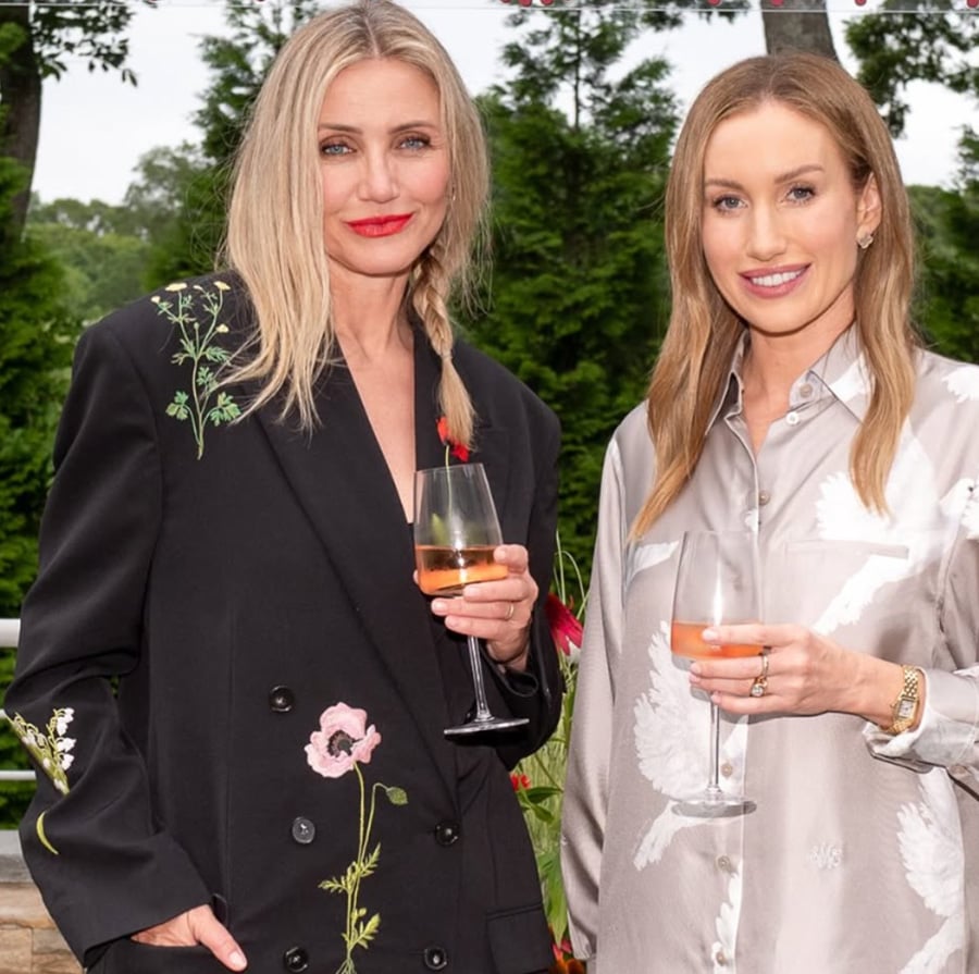 Two women standing outdoors, smiling and holding glasses of rosé wine. One wears a black jacket with floral embroidery, the other a silky light-colored shirt with white bird patterns. Green trees and plants are in the background.