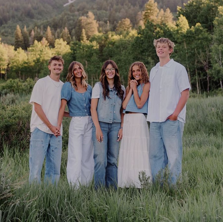 Five young people stand together in a grassy field with trees and hills in the background, smiling at the camera and wearing casual blue and white outfits.