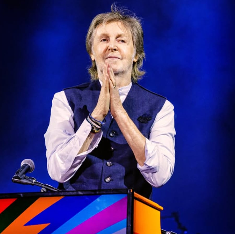 A musician with light hair stands behind a colorful piano, wearing a dark vest over a white shirt, with hands pressed together in front of his face against a blue-purple stage background.