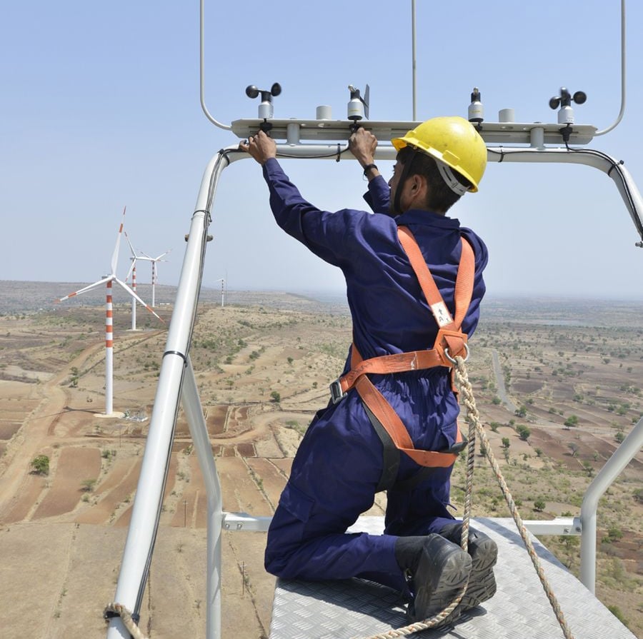 A worker in a blue uniform and yellow helmet, secured with a safety harness, adjusts equipment at the top of a wind turbine. In the background, wind turbines stand across a dry, hilly landscape under a clear sky.