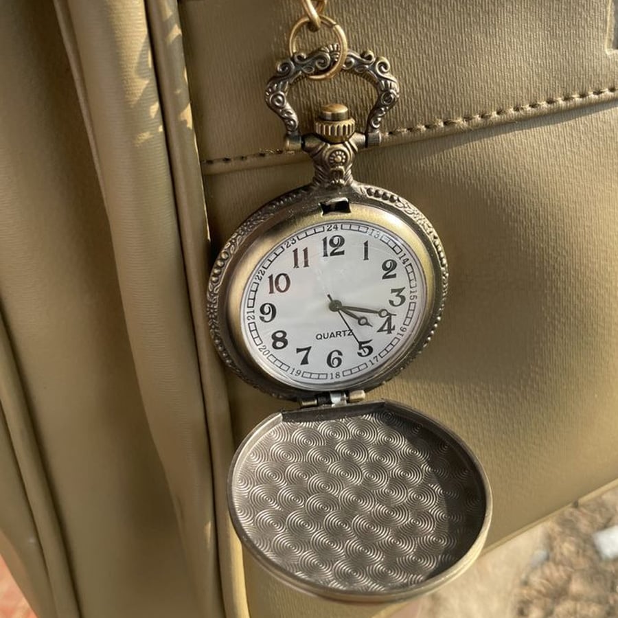 A vintage pocket watch with an ornate design hangs open, showing the time as 3:54. The watch rests against a beige textured surface, possibly a bag. Sunlight casts soft shadows on the scene.