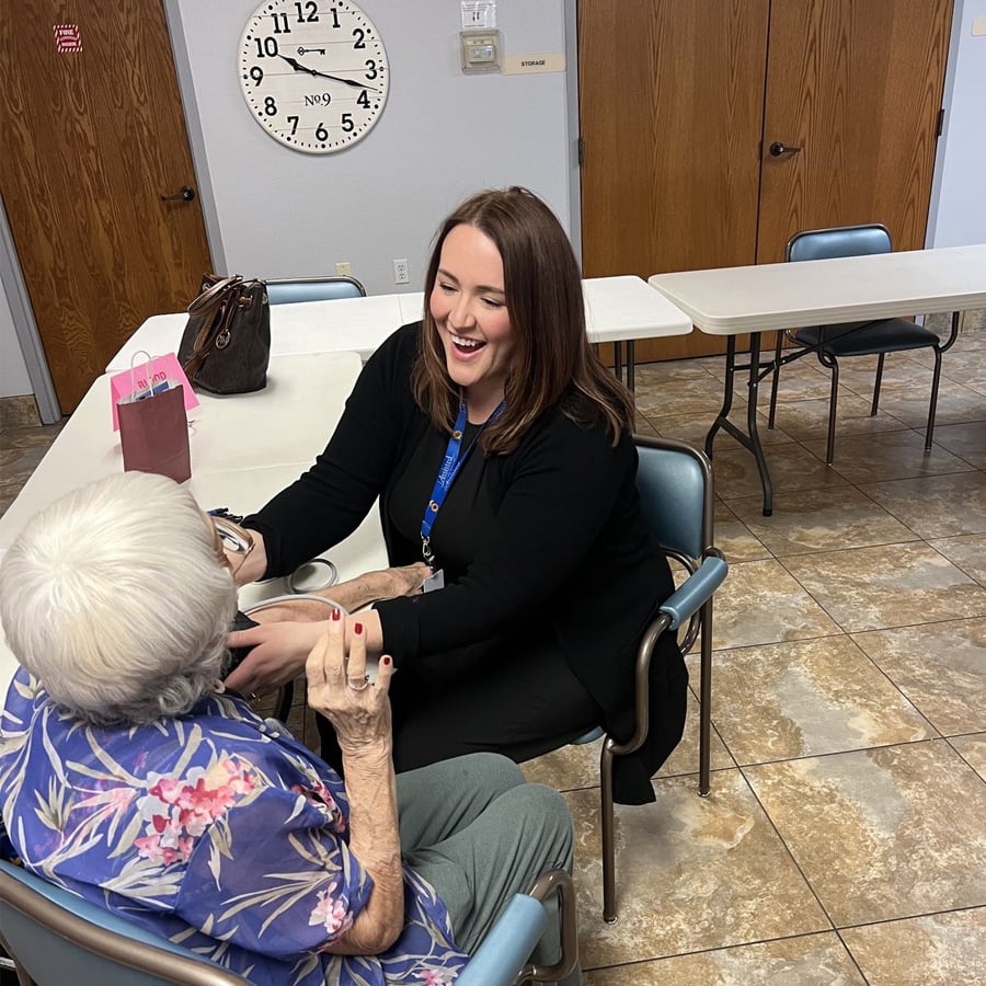 A young woman smiles and holds hands with an elderly woman in a floral shirt, sitting across from her in a room with tables, chairs, and a large clock on the wall showing 11:06.
