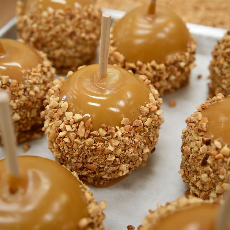Caramel apples coated with chopped nuts are arranged on a tray. Each apple has a wooden stick inserted on top for holding. The caramel is glossy and covers most of the apples.
