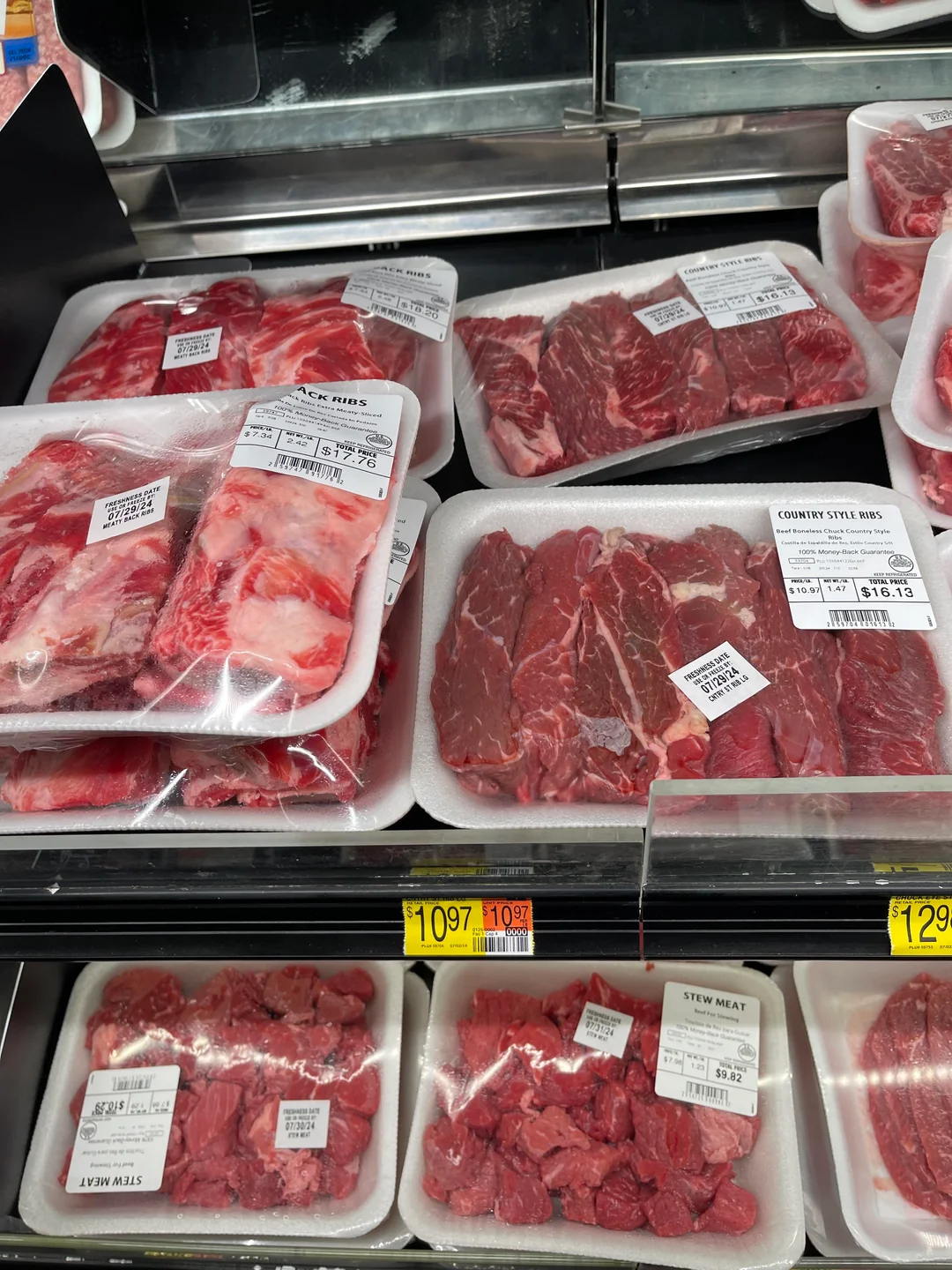 Packages of raw beef cuts, including stew meat and country-style ribs, are displayed on white foam trays wrapped in plastic in a grocery store meat section. Price tags and labels are visible on each package.