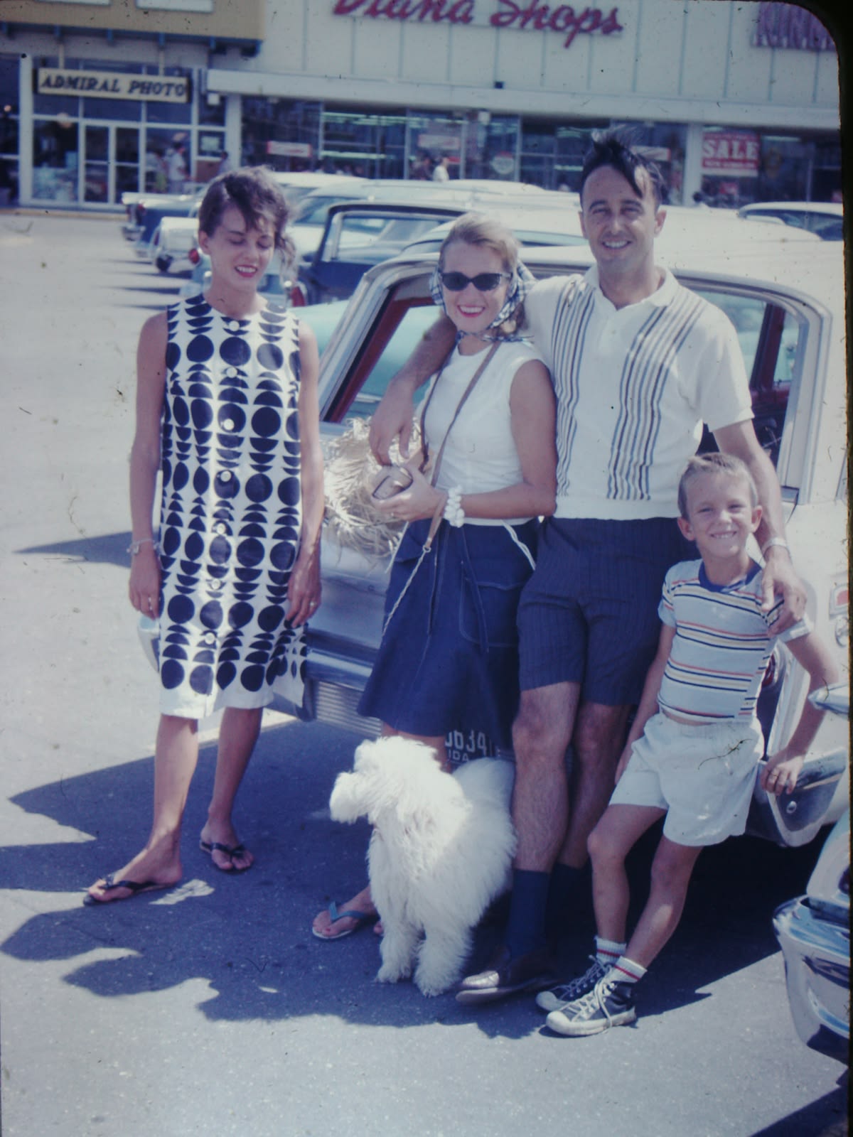 A family of four and a fluffy white dog pose by a car in a parking lot on a sunny day. Behind them are storefronts, including one called “Admiral Photo.” The family appears cheerful and casually dressed.